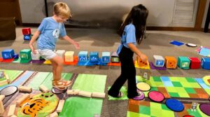 Two children walk along a story-coded floor path, following a sequence they created using large foam coding blocks. They test the code step-by-step while classmates (not visible) watch for errors to help debug the sequence.