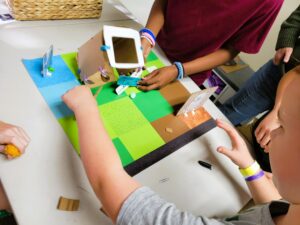 A group of children adjusts a cardboard habitat model on a table. One child secures a connector under a raised structure while others reach in to help stabilize and refine the design during a collaborative problem-solving moment.