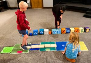 Two children walk along a tactile story-themed path they built while an adult points to large directional coding blocks, helping them follow the sequence they wrote to navigate the broken bridge and reach the destination.